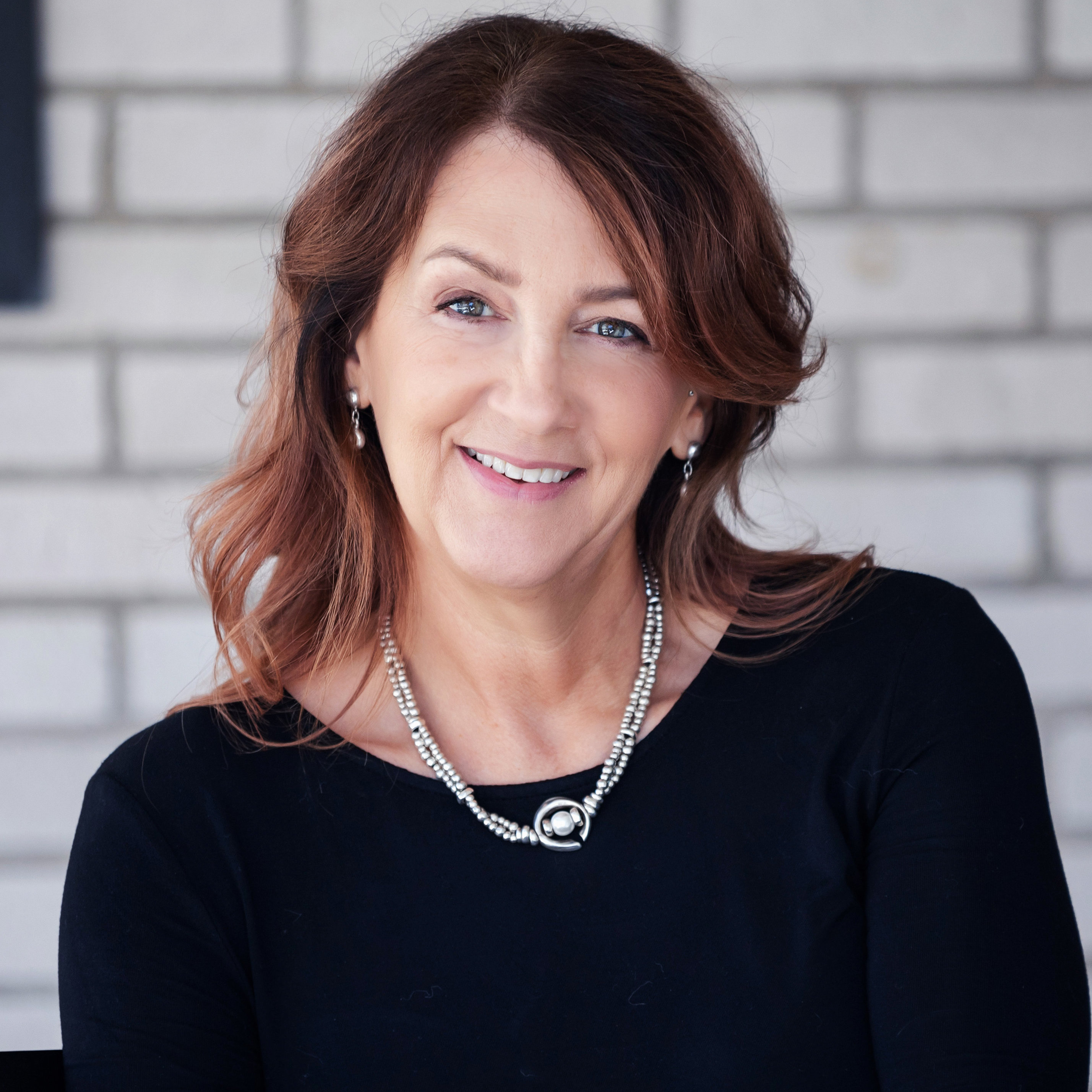 Photo: A woman in a black shirt sitting in front of a brick wall, smiling at the camera. 
