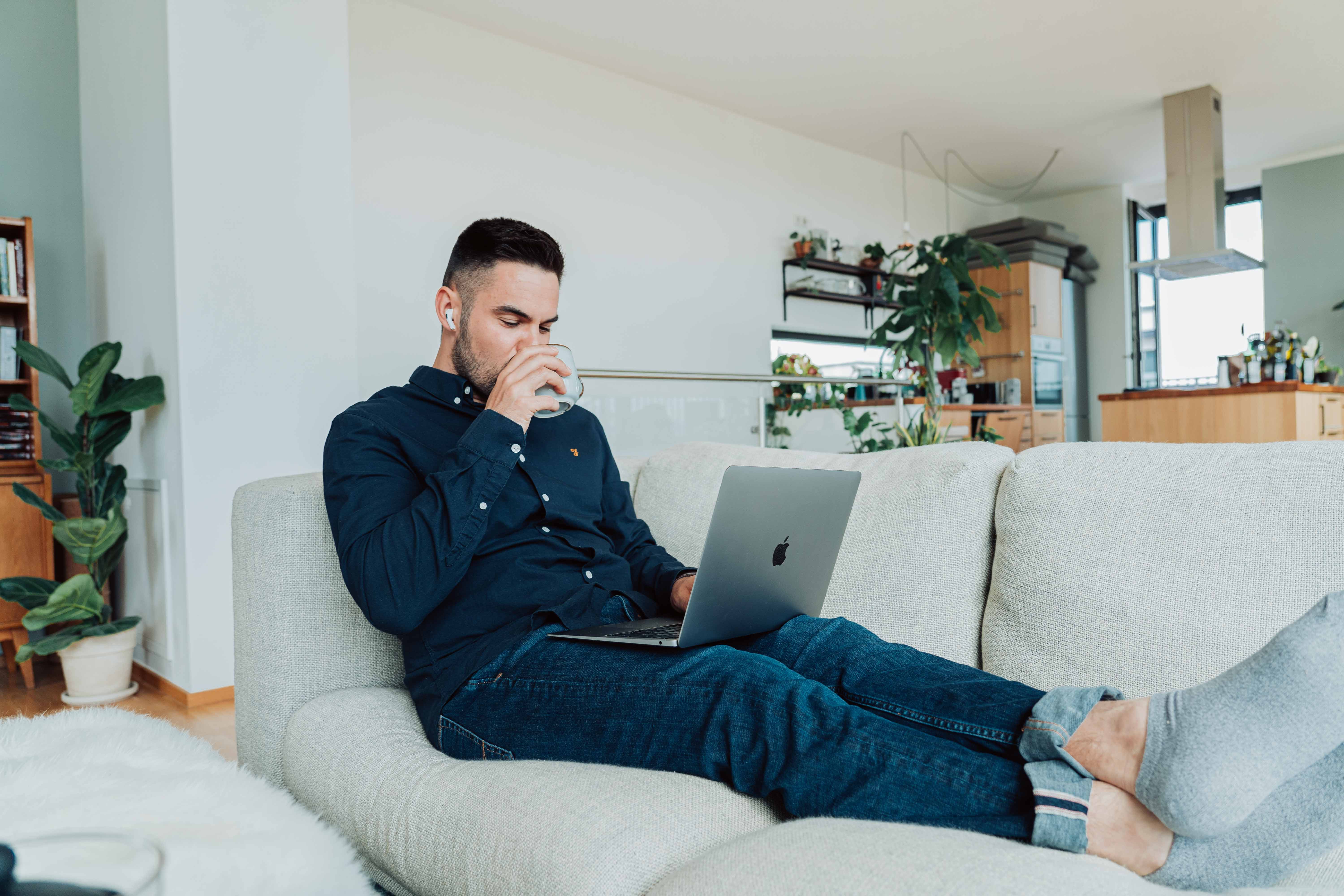 PHOTO: A white male sitting on a couch with his legs stretched out. He has a laptop in his lap and is drinking from a mug.o